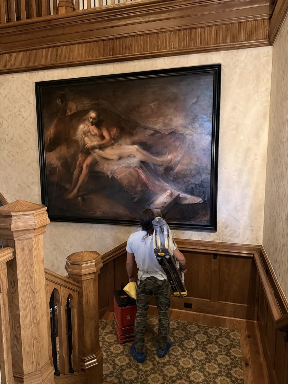 Large framed painting hung above a stair landing with oak wainscoting, with the installer standing below beside his ladder.