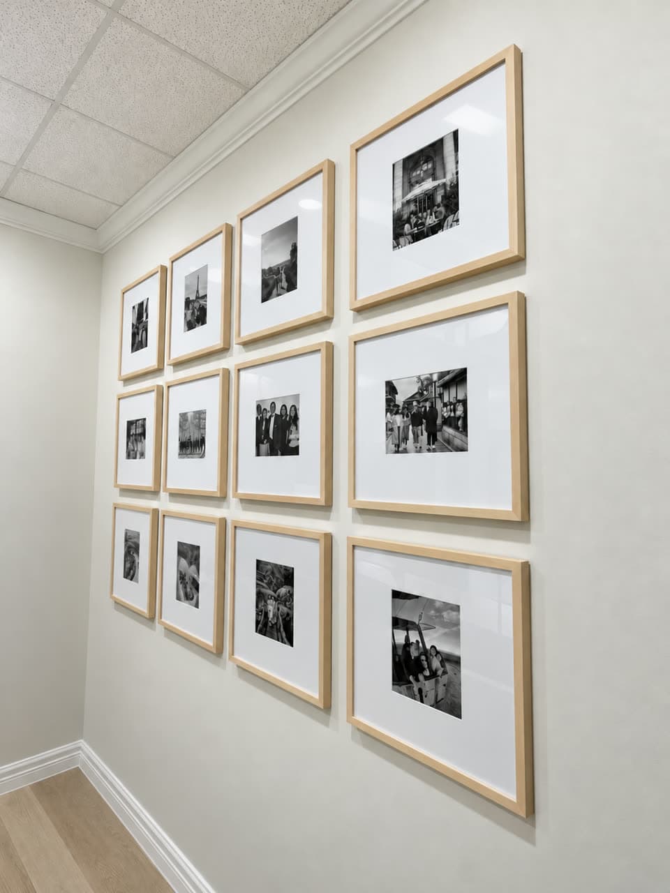 Corridor wall hung with twelve light oak frames of black-and-white photographs in an even grid.