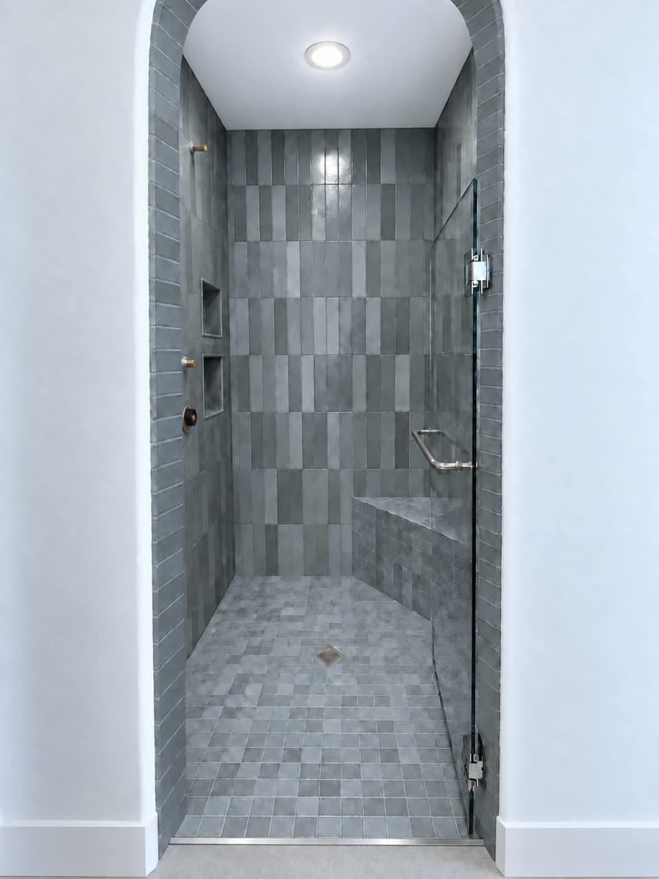 Walk-in shower behind an arched opening, tiled in grey zellige with two recessed niches, a corner bench, a mosaic floor and a glass door.