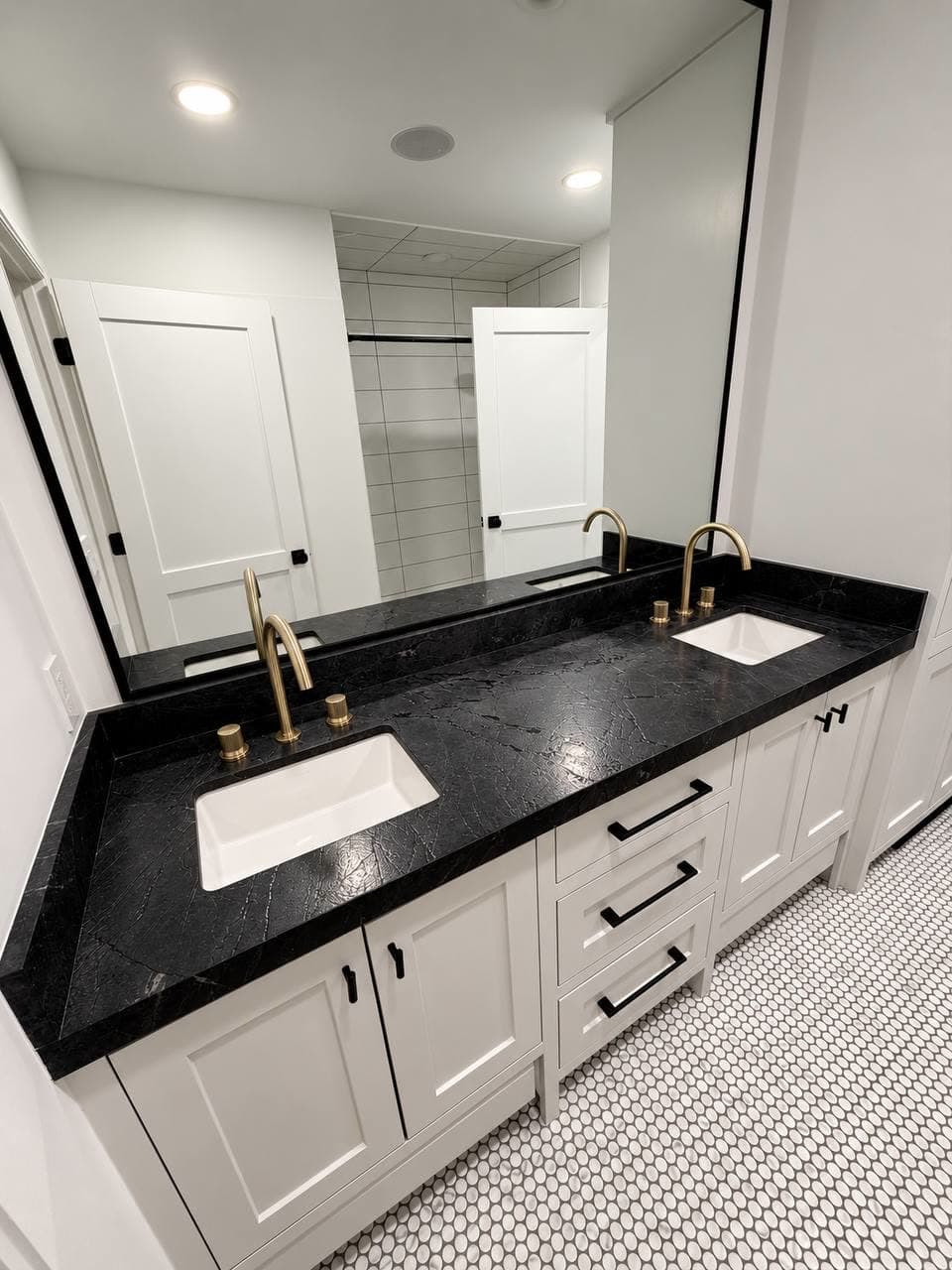Bathroom double vanity in white shaker cabinetry with a black veined stone top, brass faucets and a penny tile floor.