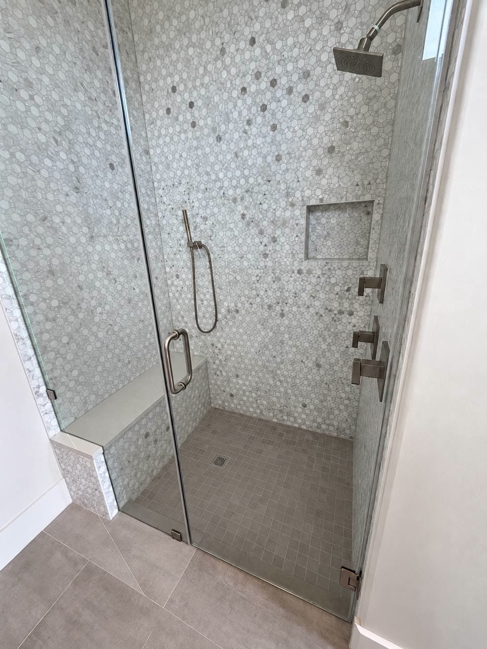 Walk-in shower tiled in grey and white hexagon mosaic with a built-in bench, a recessed niche and a frameless glass door.