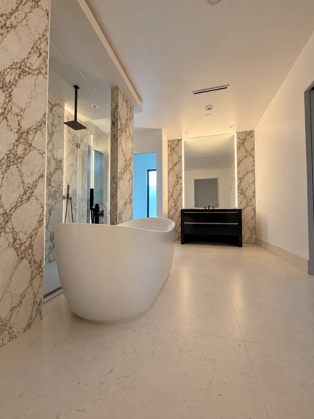 Primary bathroom with a white freestanding tub, a glass shower clad in gold-veined marble slabs, a black floating vanity between backlit marble panels and a pale large-format tile floor.
