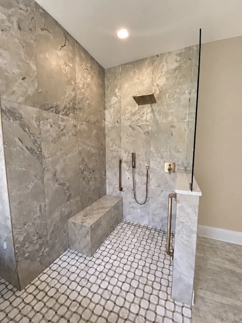 Walk-in shower in large grey stone-look tile with a tiled bench, brushed brass rain head, handshower and grab bars, a glass panel on a half wall and a marble mosaic floor.