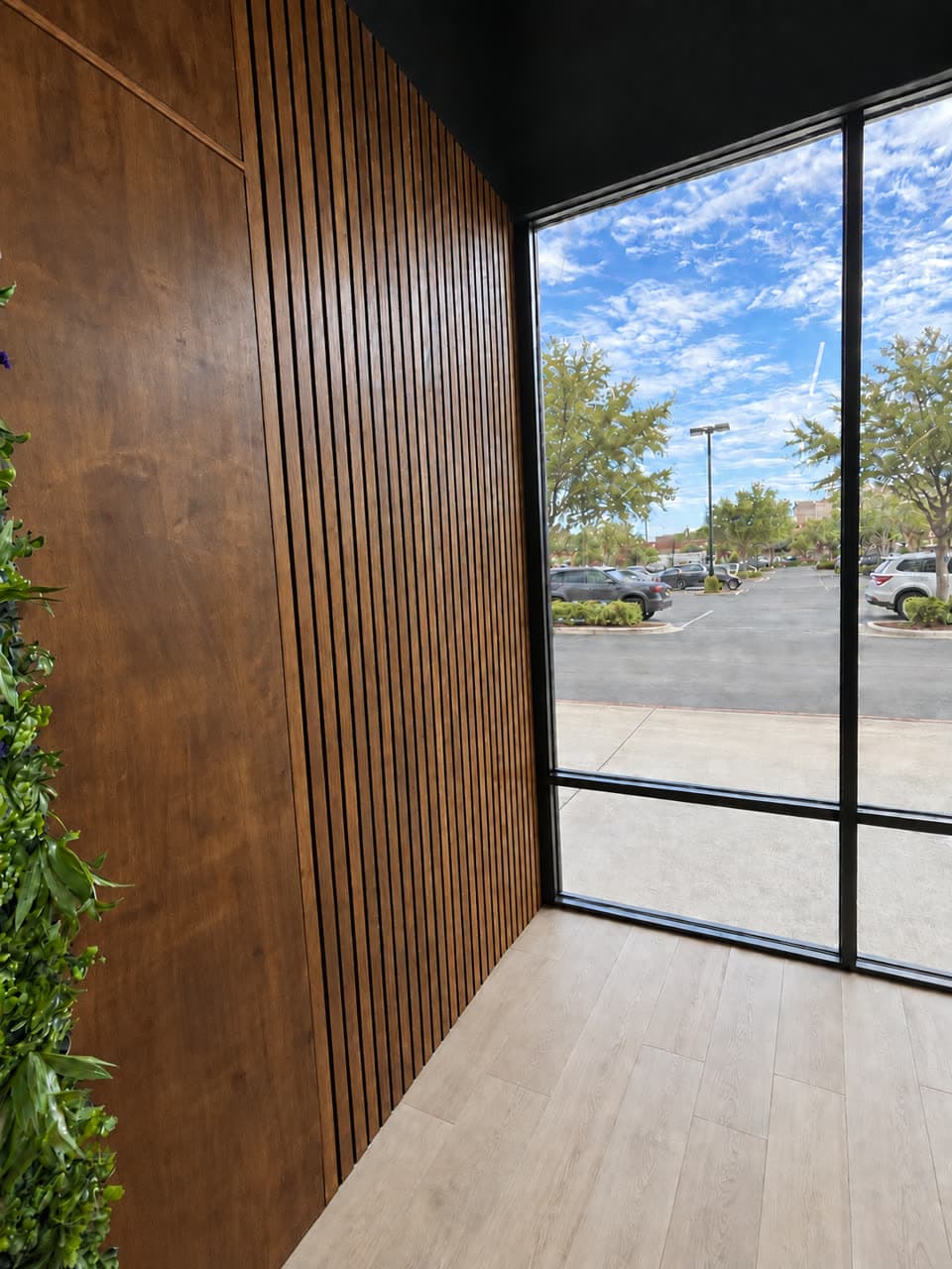 Lobby corner where a flat walnut panel wall turns into a walnut slat wall, beside a preserved moss wall and a full-height window.