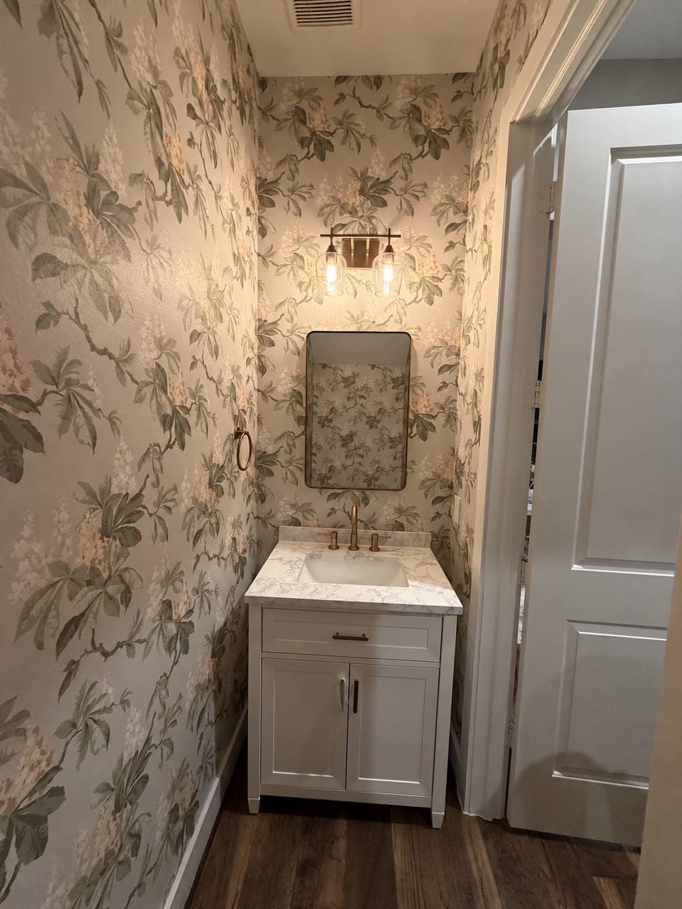 Narrow powder room papered on every wall in a soft green and blush floral, with a white vanity, a brass mirror and a two-bulb sconce.