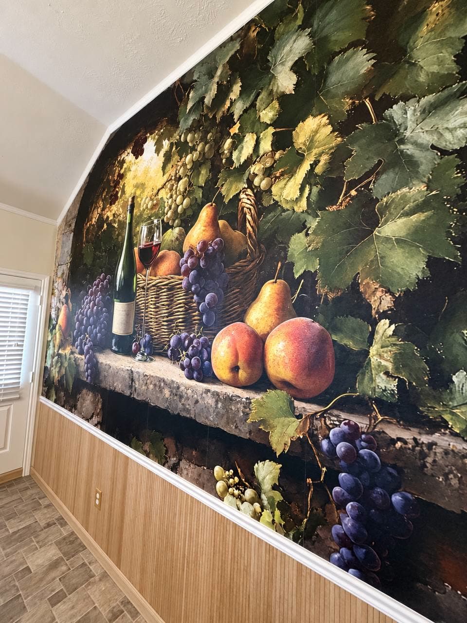 Wall covered by a painterly mural of grapes, pears and a wine bottle on a stone ledge, set above wood wainscoting.