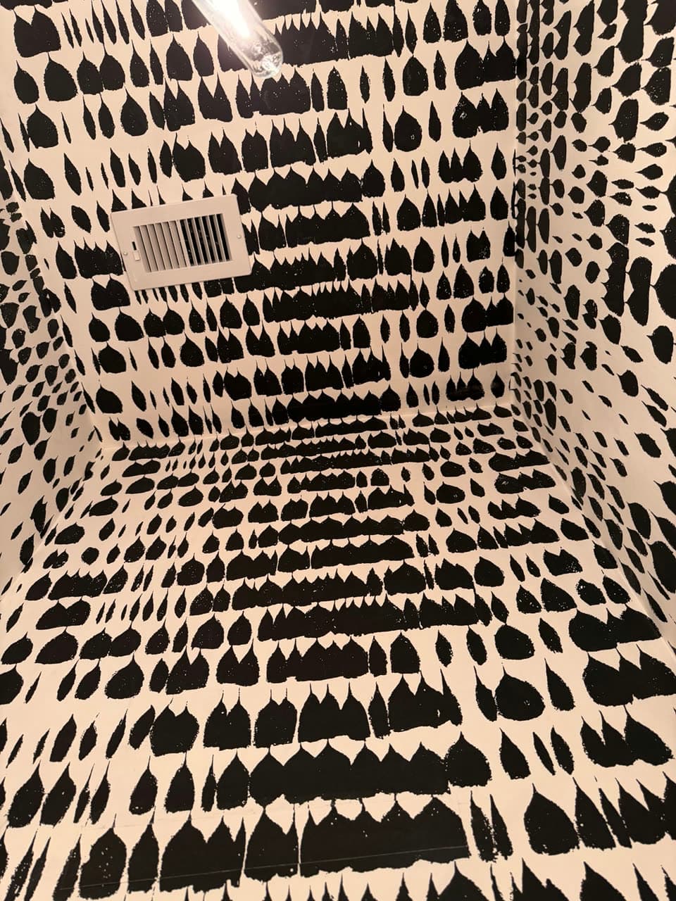 Small room papered on all four walls and the ceiling in a cream paper with black brushstroke marks, with a metal vent set into it.