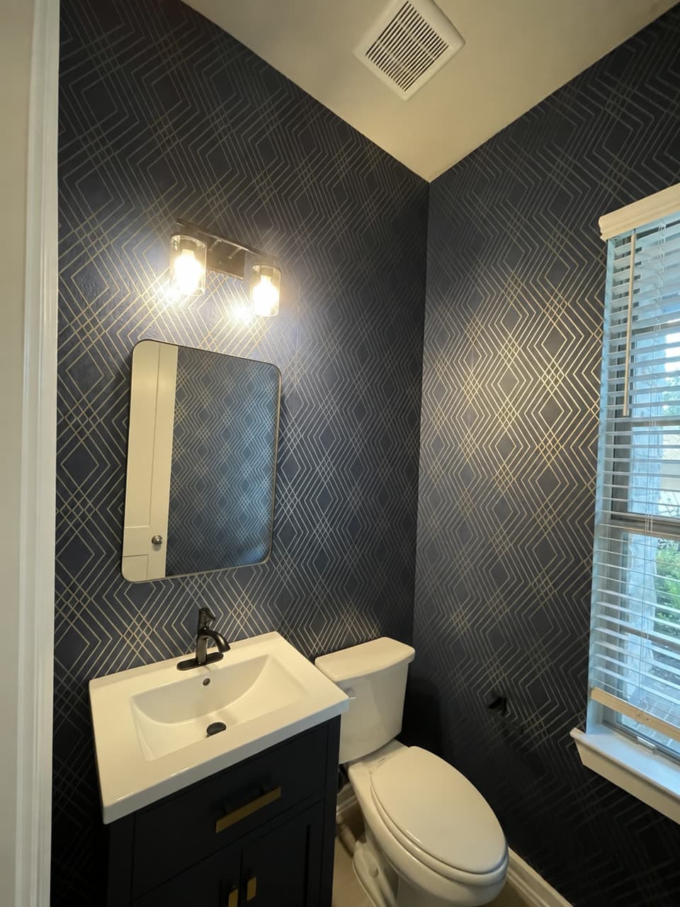 Powder room papered in a navy and gold diamond geometric, with a dark vanity, a mirror, a toilet and a blinded window.