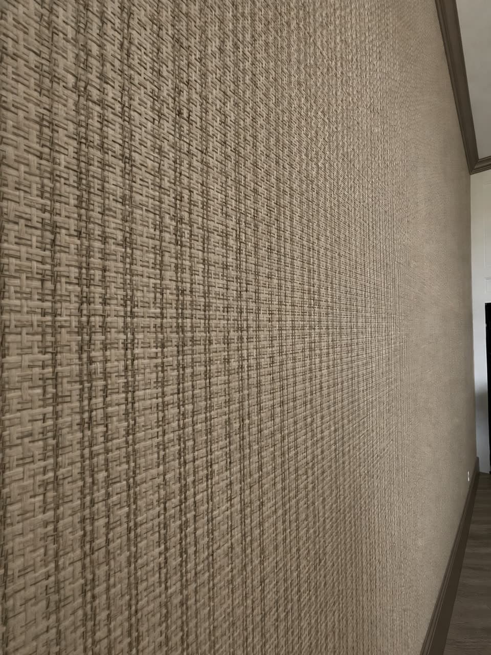 Long wall in taupe basketweave grasscloth photographed down its length, ending at dark stained trim.
