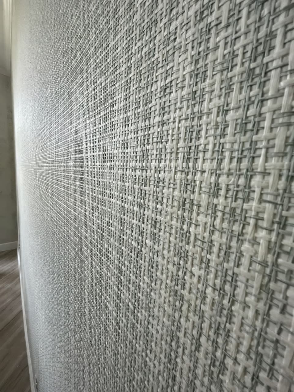 Close raking view along a wall in coarse white and green woven grasscloth, with a swept clean floor below.